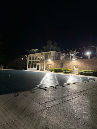 A three-story building with a white-framed glass structure at night. The building's architectural style is modern and well-lit, contrasting with the darkness. Next to the building is a paved area, reflecting the building's light. A row of hedges separates the building from the paved area. The scene evokes a sense of calmness and sophistication, accentuated by the contrast between the illuminated structure and the night sky.