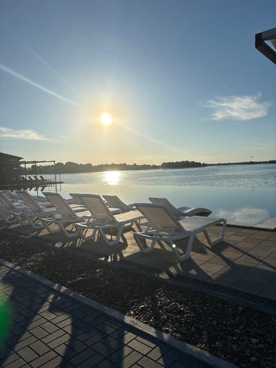 This image depicts an outdoor setting, likely a resort or a recreational area, located near the city of Kamiani Potoky, Ukraine. The scene is set during sunset, with the sun prominently visible in the upper left quadrant of the sky, casting a bright glare and long shadows. In the foreground, a row of white lounge chairs is arranged on a paved surface adjacent to a body of water, which appears to be an infinity pool or a calm lake. The water reflects the sky and the distant treeline. Beyond the water, a dense line of trees forms the horizon. In the background, a building with a dark roof and open-sided structure is visible, along with more lounge chairs. The paving stones in the foreground create a patterned walkway, bordered by a strip of gravel. The overall atmosphere is tranquil and serene, indicative of a leisure environment.
