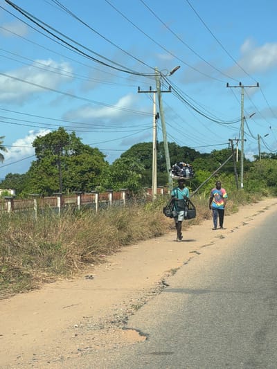 The image depicts a roadside scene in Muxara, Mozambique. Two men are walking on a dirt and concrete shoulder of a paved road. The first man is wearing a tie-dye shirt and dark pants. The second man is carrying a large bundle of clothes on his head and several bags on his body. Tall, wooden utility poles with numerous electrical wires run along the roadside. Lush green vegetation lines the edge of the road and a partially visible rusted metal fence is in the background. The sky is blue with scattered white clouds.