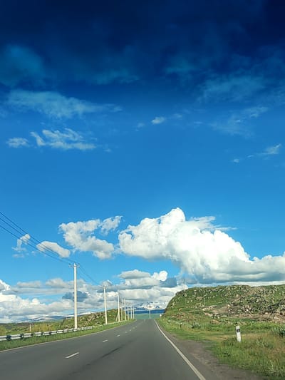 The image depicts a scenic view of a paved road curving through a rural landscape under a bright blue sky dotted with white and dark clouds. The road has clear white markings. Along the side of the road, there are utility poles with wires, a metal guardrail, and a rocky, grassy hillside. In the distance, snow-capped mountains are visible, suggesting a high-altitude location. This scene is consistent with the geography and environment of Horom, Armenia, a region known for its mountainous terrain and open roads. There are no people or animals visible in the image.