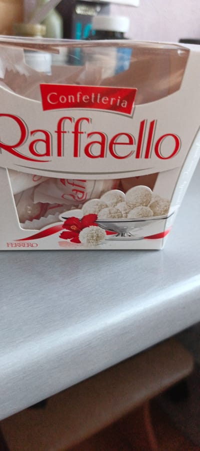 The image shows a close-up of a box of RaffaHELLO chocolates. The box is white with red lettering. The words "Confetteria" and "RaffaHELLO" are prominently displayed. Below the brand name, there are several RaffaHELLO chocolates visible, some in their wrappers and some unwrapped, sitting in a clear plastic container. There is also a red ribbon and a red flower accent on the front of the box. The box is resting on a light grey surface, which appears to be a table or countertop. The background is blurred, showing some other objects, possibly other products or packaging. There are no people in the image. The scene is indoors, and based on the provided information, it is located in the city of Tikhvin, Russia.