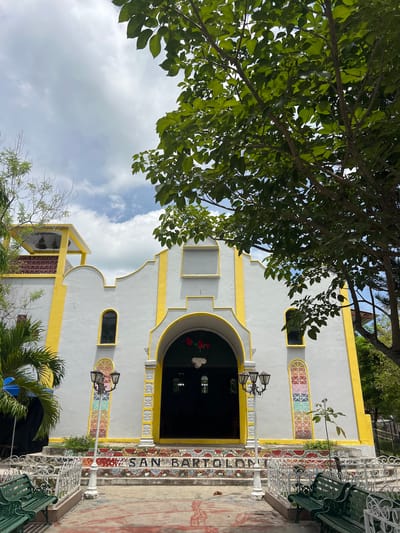 The image shows the exterior of a white church with yellow trim, located in the city of Arambala, El Salvador. The church has an arched entrance, two windows, and a bell tower. The steps leading to the entrance are decorated with colorful mosaics that spell out "SAN BARTOLON". In front of the church are two green benches, a lamppost, and some trees. The sky is cloudy.
