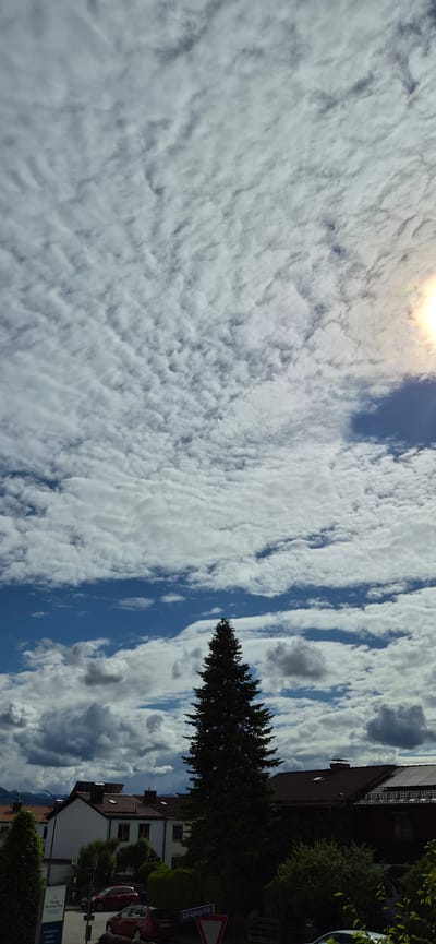 The image displays an outdoor urban landscape under a partly cloudy sky. The sky occupies the upper two-thirds of the frame, characterized by extensive altocumulus clouds forming a rippled, wave-like pattern, intermingled with patches of clear blue sky. The sun is visible in the upper right quadrant, partially obscured by clouds, generating a distinct lens flare.
A tall, dark green conifer tree, likely a spruce, stands centrally in the mid-ground, its silhouette prominent against the sky. Below the tree, several multi-story residential buildings with pitched, reddish-brown tiled roofs are visible. The building on the left features a white facade with multiple windows and is partially obscured by a white sign bearing blue text, where "Haus der..." and "Pflege" are legible. The building on the far right has solar panels installed on its roof.
In the lower portion of the image, a street is visible with parked vehicles. A red hatchback is partially visible on the left, obscured by vegetation and the aforementioned sign. A dark red station wagon is parked further right, in front of a hedge. A red-bordered triangular road sign is present near the station wagon, and a blue street sign attached to a pole clearly displays "Josef-Ramberger-Straße," unequivocally locating the scene in Kolbermoor, Germany. Hedges and various smaller trees and bushes are distributed throughout the foreground and around the buildings. Faint outlines of mountains are discernible on the distant horizon, particularly towards the left. No people are visible within the frame.
