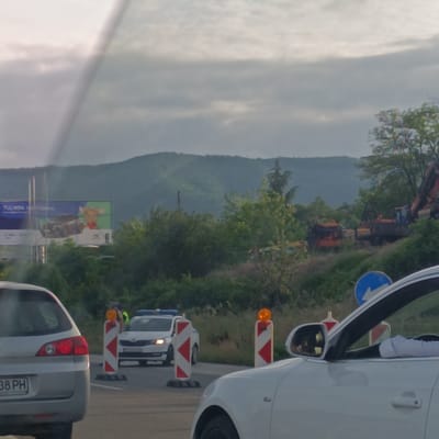 The image depicts a scene in Dolno Varshilo, Bulgaria, possibly near a highway construction or traffic control area. In the foreground, the rear of a silver car is visible on the left, with a license plate reading "38 PH". To the right, the front portion of a white car is seen, with a side mirror reflecting a distorted view.

In the middle ground, a police checkpoint is set up. A white police car with flashing blue lights is positioned behind two red and white striped traffic cones, each topped with an orange flashing light. A police officer, wearing a high-visibility vest over a uniform, is standing near the police car, interacting with someone. A blue and white directional sign is also present in this area.

Further in the background, several construction vehicles are visible, including what appears to be an excavator. The landscape features lush green trees and rolling hills under a cloudy sky. A billboard with advertisements for "Tucson GO" and a cartoon character is also visible on the left side of the image, suggesting the location is near a main road.