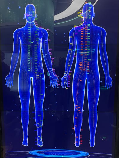 Located in Guangming District, China, the image displays a large, vertically oriented digital screen featuring two glowing blue wireframe human anatomical models against a dark blue background. The model on the left presents an anterior view, while the model on the right presents a posterior view. Both figures are overlaid with numerous small colored labels, predominantly in white, green, yellow, pink, and red, depicting Chinese characters that correspond to specific acupuncture points and meridian lines. Thin, faint lines connect these labels to the wireframe bodies. The background exhibits subtle digital patterns, including faint vertical lines resembling code on the upper left and scattered luminous dots. Reflections on the screen surface reveal partial figures of individuals and elements of the surrounding environment; a discernible reflection of a person, possibly holding a camera, is visible on the right side of the display, overlapping the lower back of the right anatomical figure. A faint, glowing blue ring pattern is situated at the base of both figures. No direct human subjects are present within the primary image content; only reflections are observed.