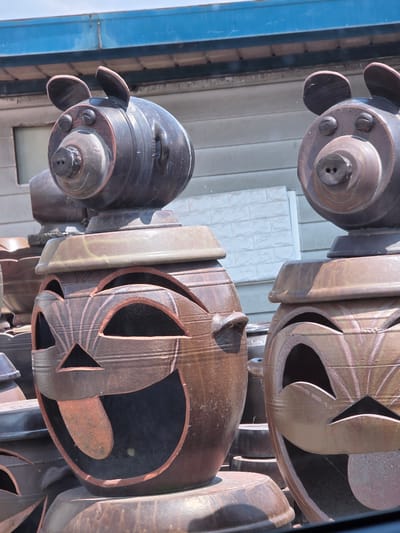 The image shows a close-up of several large, decorative ceramic jars, likely for sale as garden or decorative items. The foreground features two jars with carved faces that resemble jack-o'-lanterns, with wide, open mouths and triangular eyes, and one with a prominent tongue sticking out. These jars are stacked in rows, with parts of other similar jars visible behind them. On top of the closest carved jar, a smaller ceramic pig head is mounted. Another pig head is partially visible on a jar to the right. The jars are a mottled brown and reddish-brown color, with a glazed finish. The background shows a blue corrugated metal overhang and a light-colored textured wall, suggesting an outdoor market or workshop setting. The scene appears to be located in Cheongju-si, South Korea, a city known for its pottery. There are no people visible in the image.