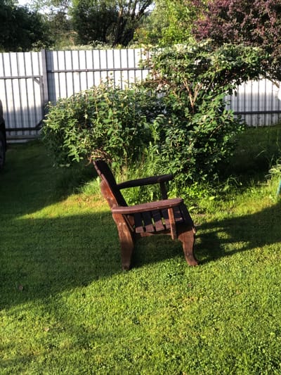In Staroe Istomino, Russia, a wooden Adirondack chair sits on a vibrant green lawn in front of a white corrugated metal fence. Lush green bushes and trees with some darker foliage are visible behind the fence and around the chair, casting dappled shadows on the grass. The sun illuminates the scene, highlighting the textures of the lawn and the chair.