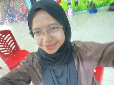A portrait-oriented, slightly blurry selfie features a woman wearing a black hijab and glasses, looking towards the camera with a slight smile. She is wearing a brown outer garment over a dark inner garment. To her left, a red plastic chair with a distinctive slotted design is visible. In the background, the environment appears to be indoors, possibly a classroom or play area, with colorful wall decorations and what appear to be children's bags or cushions on the floor. The floor is tiled and light-colored. There are no overt indications that this scene is specifically in Tanjung Gading, Indonesia, beyond the prompt's assertion.