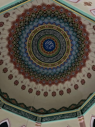 The image shows an ornate dome ceiling, likely inside a mosque. A central circular design features a blue medallion inscribed with Arabic calligraphy. Concentric rings radiate outwards, patterned with alternating colours and geometric motifs. The dome's curvature is accentuated by floral-like patterns extending towards the edges. The perimeter showcases further calligraphy along the architectural framework, with a combination of pale hues and light green shades providing a soft backdrop.