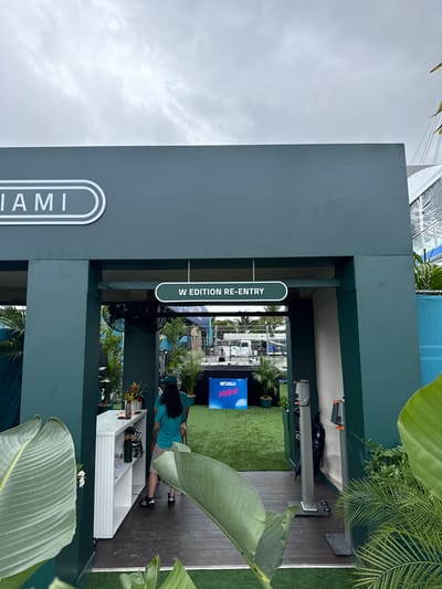 Here is a detailed description of the image:
The image shows an entrance at an event in Miami Gardens, United States. A large sign on the left reads "MIAMI" vertically. Above the entrance, another sign reads "W EDITION RE-ENTRY," indicating it is a re-entry point for the "W Edition" event. The structure surrounding the entrance is a dark green color.
Inside the entrance, there is artificial green turf. A television screen displays "MIAMI" with pink and blue accents. There is a person with dark hair wearing a blue shirt and shorts working at a white counter. The counter has several items on it.
The image is taken outdoors under an overcast sky. Lush tropical foliage is visible in the foreground and around the entrance, enhancing the Miami atmosphere. The overall scene is clean and well-maintained, suggesting a professional and upscale event.