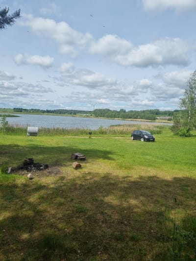 The image shows a serene landscape in the vicinity of Kalniškiai, Lithuania, featuring a body of water, a grassy field, and a clear sky with scattered clouds. In the foreground, a campfire pit with remnants of burnt wood is visible, along with some logs and a rock. Further back, a black car is parked on the grass, and a metallic structure resembling a bin or a small shed stands near the water's edge. The water is calm, reflecting the sky, and bordered by reeds and trees. In the distance, a wind turbine and some buildings can be seen, suggesting the presence of a rural or semi-rural community. The overall scene evokes a sense of peace and natural beauty, characteristic of the Lithuanian countryside.
