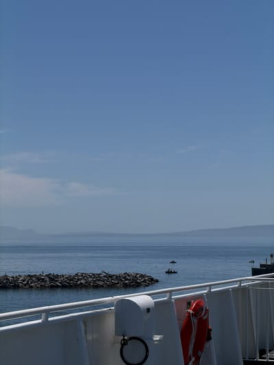 The image depicts a view from a ferry in Delta, Canada, looking out towards the water. In the foreground, the white railing of the ferry is visible, along with a red life preserver. Further out on the water, a rocky breakwater juts into the sea, and several small boats with people can be seen. In the distance, hazy mountains form the horizon under a clear blue sky.