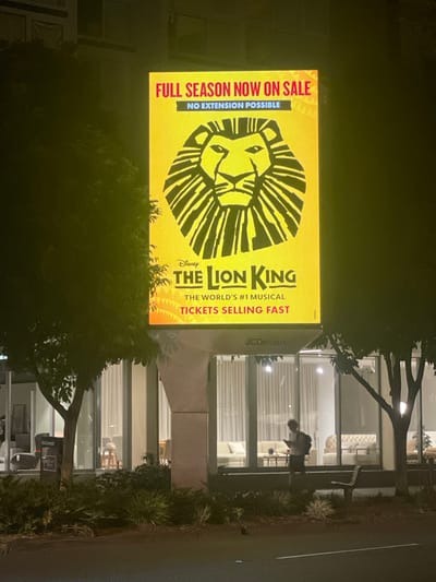 The image shows a large, illuminated billboard for Disney's "The Lion King" musical. The billboard features the iconic lion's head logo and text that reads "FULL SEASON NOW ON SALE NO EXTENSION POSSIBLE THE WORLD'S #1 MUSICAL TICKETS SELLING FAST." The billboard is positioned above a building with large windows, displaying indoor furniture. To the left of the billboard is a tree, and to the right is another tree with a silhouette of a person standing in front of the building. The scene appears to be at night, as indicated by the lighting and the dark environment. Based on the presence of the JCDecaux advertisement panel on the lower part of the billboard structure, this scene is likely located in a public urban space. The architecture of the building and the type of billboard suggest a commercial or entertainment district. Without further contextual information or specific landmarks visible, pinpointing the exact location within Brisbane, Australia is not possible from the image alone.