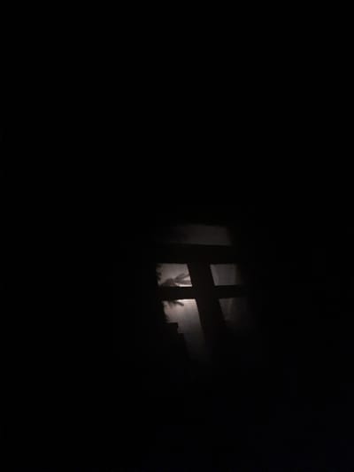 The image shows a dark scene with a window frame in the center, silhouetted against a faint glow. The window frame has a cross shape. Through the window, a blurry, pale circular object, possibly the moon, is visible. The overall impression is of a nighttime view through a window, with little detail discernible due to the darkness. There is no discernible location in the city of Liubakhy, Ukraine, or any people, actions, or interactions.