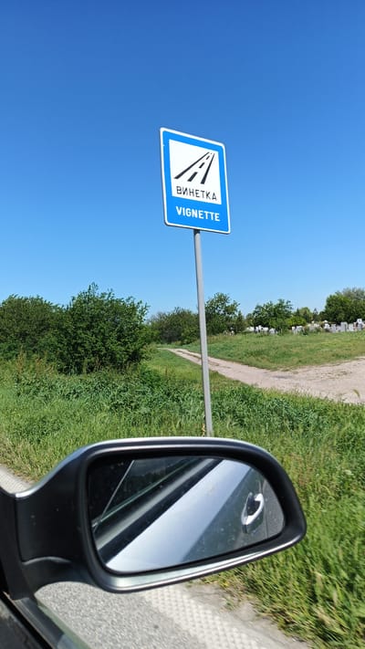 The image depicts a roadside scene, likely taken from inside a moving vehicle. A road sign indicating "Vignette" is prominently displayed against a clear blue sky. The sign features a stylized image of a highway. The foreground includes the side mirror of a car, partially obstructing the view. The background shows a grassy field with a dirt path leading towards a cemetery or similar area marked by rows of white or light-colored structures. Lush green vegetation is present on both sides of the road. The location is the city of Boliartsi, Bulgaria. No people are visible in the image.