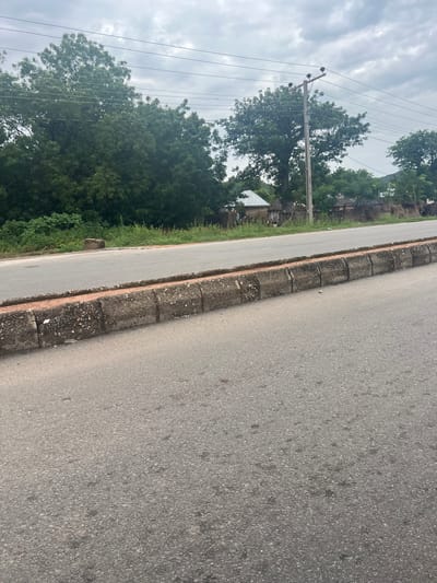 The image depicts a roadside scene in Kona Garu, Nigeria. The perspective is low and angled, looking along the edge of an asphalt road. The road itself is the most prominent object, with a textured grey surface. To the left of the road is a concrete curb, also textured and showing signs of wear. Beyond the curb, there is a strip of green grass and vegetation, which then gives way to a dense collection of trees. Some of these trees are quite large with lush green foliage.
In the background, through the trees, glimpses of buildings can be seen, suggesting a semi-rural or residential area. A prominent feature is a utility pole with several wires stretching across the frame. The sky is overcast, with a cloudy, grey appearance, indicating a potentially humid or rainy day. The lighting is diffused, suggesting it is daytime, but the lack of strong shadows prevents a precise time of day from being determined. There are no people visible in the image. The overall mood is neutral, capturing a common roadside view. No text is visible in the image.