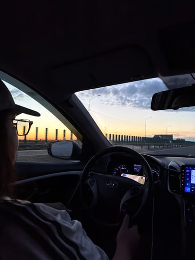 A view from inside a car driving along a road, potentially a bridge, at dusk or dawn. The driver is holding the steering wheel. The car's dashboard and infotainment system are visible. Through the windshield and side windows, the sky is lit with the colors of the sunrise or sunset, reflecting off the glass. A pair of eyeglasses rests on the dashboard in front of the passenger seat. Based on the map visible on the car's screen, the odometer reading, and other car details, it is difficult to pinpoint the exact location in Kislyakovskaya, Russia.
