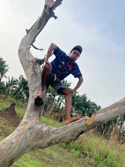 An eye-level photograph taken in Agba Umana, Nigeria, shows a young man sitting in the fork of a large, dead, light-colored tree trunk with a prominent branch extending towards the left. The man is wearing a short-sleeved, dark-blue and white striped soccer shirt with a logo, dark shorts, and a dark baseball cap. He is barefoot and his skin tone is dark. He is posed with his right hand on his right knee and his left hand on his hip. The background consists of a cloudy sky, tall green grass, and leafy trees. The tree itself has a large break in the bark visible on the right side of the frame. The ground below the tree is partially visible and appears to be grassy and slightly damp.