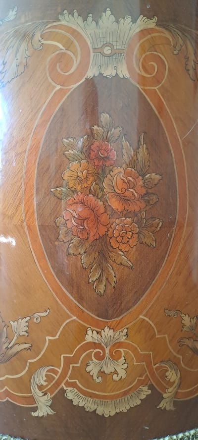 The image shows a close-up view of an ornate wooden surface, likely a piece of furniture, decorated with marquetry and painted floral motifs. The central element is an oval-shaped inlay featuring a bouquet of vibrant orange and yellow flowers with green and brown leaves, rendered in a painterly style. This oval is framed by concentric borders of different wood tones and inlaid details, including scrollwork and decorative elements in lighter wood and a reddish-brown hue. The background wood grain varies in color and direction, creating a textured and visually rich surface. The style of the decoration suggests an antique or vintage piece. The image is a photograph taken indoors, likely of an object within a home environment. There is no indication of a specific location in Hässelby, Sweden.