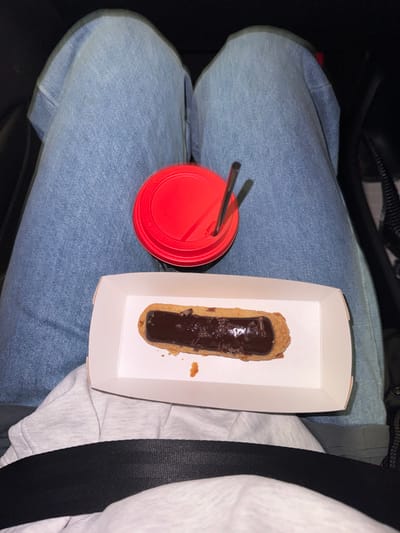 A red-lidded cup with two straws rests on blue jeans, next to a white cardboard container holding a chocolate éclair. A gray seatbelt strap lies adjacent to the container. The background is dark, hinting at a car interior.