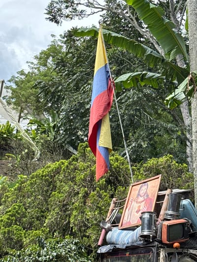 The image depicts an outdoor scene in Quimbaya, Colombia, under an overcast sky. A Colombian flag, oriented vertically on a pole, hangs prominently in the upper central portion of the frame, displaying its yellow, blue, and red bands. Beneath the flag, on what appears to be the roof or cargo area of a vehicle, a collection of items is secured. These objects include a framed religious print of Jesus Christ, two metallic coffee percolators, a small orange vintage television set, and a metal ladle, alongside other unidentifiable items and folded textiles. The background consists of dense, lush green vegetation, featuring large banana leaves and various species of trees and shrubs. No individuals are present.