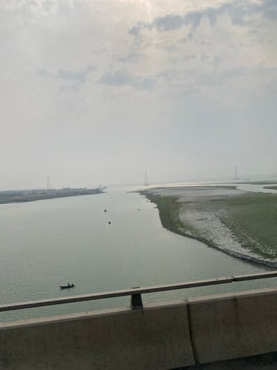 The image depicts a wide river landscape in Naodoba, Bangladesh, captured from an elevated perspective, likely a bridge. In the immediate foreground, a concrete bridge barrier with a metal railing extends horizontally across the bottom.

The primary subject is a broad, greenish-gray river, likely the Padma River, flowing through the scene. On the river, one dark-hulled boat carrying at least two individuals is visible in the lower-left midground. Further upstream, two smaller, indistinct dark boats appear as specks on the water.

To the left, a vegetated landmass or island stretches into the distance, with visible signs of development or industry, including a crane-like structure and an embankment at its far end. To the right, a large expanse of mudflats and low-lying land, sparsely covered with green vegetation, forms a curved bank along the river's edge, indicating exposed riverbed.

In the background, three prominent lattice-style electrical transmission towers stand tall, two appearing on or near landmasses within the river's expanse and one slightly further right on the distant right bank. The sky is overcast with diffuse sunlight penetrating the clouds, creating a bright, hazy atmospheric condition.
