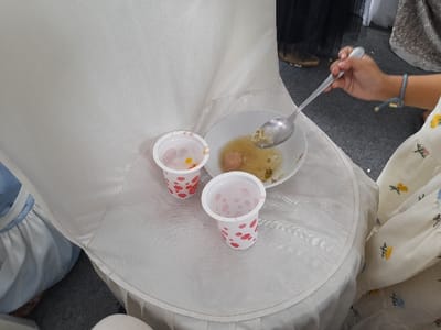 The image captures a close-up view of a person's hand holding a spoon, eating from a bowl of soup. The bowl, containing noodles and meatballs in a light broth, rests on the white fabric slipcover of a chair. Beside the bowl are two disposable plastic cups; one holds colorful, small, jelly-like items, and the other is empty. The person eating wears a white dress adorned with embroidered flowers, and a blue and grey bracelet is visible on their wrist. In the background, parts of a person in dark clothing are visible, standing on a grey carpeted floor. The scene suggests an indoor gathering or event, possibly a casual meal during a celebration, consistent with the provided location context of Kecamatan Gempol, Indonesia. There is no visible text in the image.