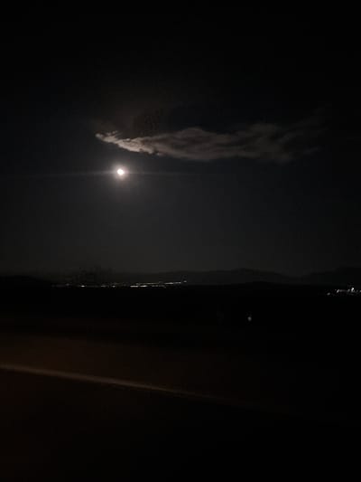 The image appears to be taken at night. A bright light source, likely the moon, is partially obscured by a cloud. To the right of the light source is a dark, vertical structure that could be a building or a large object. The overall environment is dark and it is difficult to make out details. There are no visible people, actions, or interactions in the image. Given the prompt mentioning the location, the picture could be taken in Lesichovo, Bulgaria.