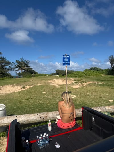 The image depicts a woman in a bikini and wrap sitting in the back of a pickup truck at Hukilau Beach Park in Laie, United States. The woman has blonde hair and is facing away from the camera towards a blue park sign indicating "HUKLAU BEACH PARK". The sign lists regulations prohibiting alcoholic beverages, drugs, loitering, overnight parking, and soliciting. The pickup truck bed contains several water bottles and a drink. The environment includes grass, trees, sand, and a blue sky with clouds.