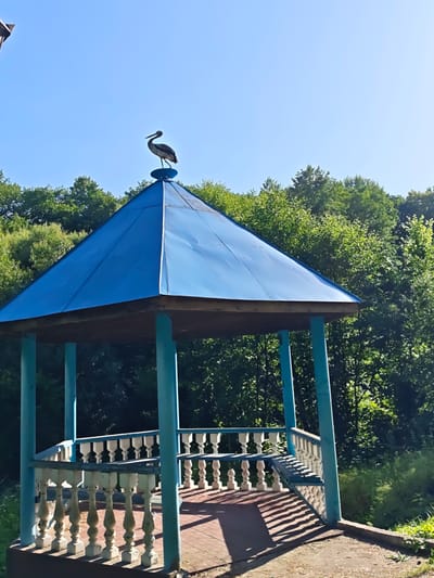 A blue gazebo with a metal roof and white balusters stands in a wooded area. A silhouette of a bird is perched on top of the gazebo's roof. The gazebo is located in the city of Sosny, Ukraine. The surrounding environment consists of lush green trees and foliage, under a clear blue sky. The ground beneath the gazebo is paved with reddish brick.