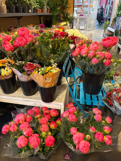 A horizontal, high-angle shot displays a selection of flowers in a flower shop, presumably in Riedlingen, Germany. Buckets of pink peonies, wrapped in clear plastic, dominate the foreground and upper-right corner. Other flower arrangements in buckets are visible on a lower shelf. A blue, multi-tiered display stand holds additional bouquets. In the background, a hair salon with a stylist and client is seen through a glass entrance. Shelves of products and decorative plants are visible on the left side.
