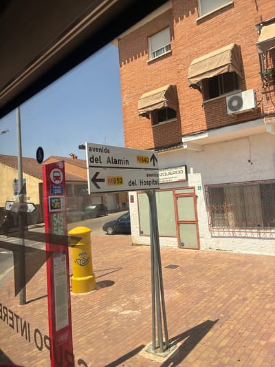 This is a ground-level exterior shot of a street in Villa del Prado, Spain, likely taken from inside a vehicle due to the reflection on the left side of the image. The scene is dominated by a brick building with awnings over the windows and an air conditioning unit mounted on the facade. In the foreground, a red bus stop sign displays route information and a bus icon, next to a yellow mailbox with a post office emblem. Two directional street signs are visible; one points right with "avenida del Alamin M540" and an upward arrow, and another points left with "M952 del Hospital" and an arrow. A white building with a business sign for " Condiciones Electricas" is also present. The ground is paved with reddish-brown bricks laid in a patterned arrangement. The sky is clear and blue. Several vehicles are parked or driving on the street in the background.