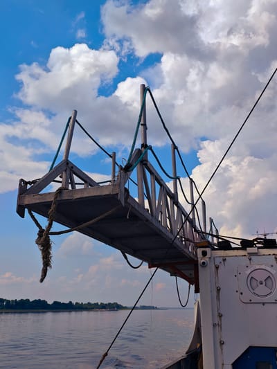 A metal gangplank extends from a vessel onto a calm body of water under a partly cloudy blue sky. The gangplank is supported by ropes and metal struts and appears weathered. The side of the vessel is white with visible bolts and a circular gauge. In the distance, a tree-lined shore is visible, and on the water, a barge is present. The overall scene suggests a river or harbor environment. The location is Erkalovo, Russia.