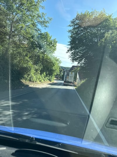The image shows a view from inside a vehicle, looking out through the windshield. The scene depicts a road with trees lining one side and buildings in the distance. A white box truck is visible on the road, with some text on its side. A person is standing near the back of the truck. The sky is blue with some clouds, suggesting it is daytime with good weather. The presence of trees and a road indicates a rural or suburban setting. The image is captured from a slightly angled perspective, possibly as the vehicle is in motion.
