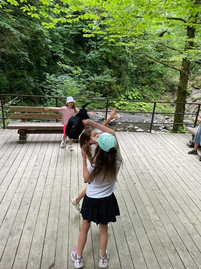 Two young girls are interacting on a wooden deck overlooking a natural landscape in Izmaylovka, Russia. The girl on the left, wearing a pink t-shirt, white cap, and shorts, is standing near a wooden bench. She has a black backpack slung over her shoulder and is looking towards the viewer with a smile. The girl on the right, wearing a white t-shirt, black polka-dot skirt, and a light blue cap, is in the foreground, leaning back slightly and touching her head with one hand. Her long brown hair is visible. She is also wearing sneakers. The deck is made of weathered wooden planks. Beyond the deck, lush green trees and foliage cover a hillside, with a small stream or rocky creek visible in the background. The scene appears to be set during the daytime, with natural light illuminating the area. The overall mood seems lighthearted and playful. There is no visible text in the image.