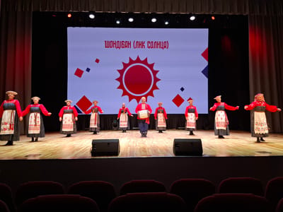 The image shows a stage performance in Kungur, Russia. A group of ten performers, dressed in traditional red and white embroidered costumes, are positioned on a wooden stage. Nine of the performers are women, and one in the center is a man holding a drum. They are all facing forward, with some of the women extending their arms. A large screen behind them displays a graphic of a red sun and text in Russian that translates to "Shondyban (Face of the Sun)." Two black speakers are placed on the stage floor at the front. The stage is lit by spotlights, and red curtains frame the sides of the screen. In the foreground, rows of dark red theater seats are visible, suggesting an audience is present or has been seated. The overall scene depicts a cultural performance taking place indoors.