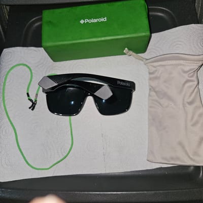 The scene depicts a pair of black Polaroid sunglasses with a green lanyard resting on a white paper towel. Next to the sunglasses is a green Polaroid box and a beige fabric pouch, also likely for the sunglasses. The items are arranged inside a black tray or container. There is no information to determine the location is Vic, Spain.