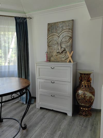 This media file depicts an indoor scene, likely a room in a home, with elements suggesting a decorative and perhaps spiritual aesthetic. The main subjects are decorative objects arranged in a room with white walls and light gray wood-plank flooring.
Prominently featured is a large, textured wall hanging of a Buddha face, rendered in muted earth tones. To the right of the Buddha head, on top of a white, three-drawer dresser, sits a light-colored wooden letter "X". Also on the dresser, to the left of the "X", is a small, pink, mannequin-shaped jewelry holder. Next to the dresser, to its right, is a very large, ornate vase with intricate patterns in gold, red, and blue, depicting floral and possibly animal motifs.
To the left of the dresser and Buddha head is a window draped with sheer white curtains and heavier, dark blue velvet curtains. In the foreground, to the left of the frame, is part of a round wooden table with a dark metal, ornate base.
The lighting suggests daytime, with natural light filtering through the window. There are no people visible, and no specific activity is taking place; it is a still-life arrangement of decor. The overall impression is one of a thoughtfully decorated interior space. The location context provided indicates this scene is from Квант-88 Снт, Russia.