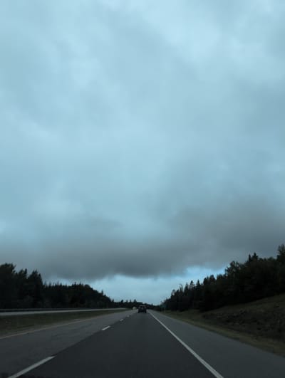 The image shows a highway in Quispamsis, Canada, under a cloudy sky. The road is asphalt with white and yellow lane markings. Trees line the right side of the road, forming a dense forest. A dark-colored car is visible in the distance on the right side of the road, and a lighter-colored car is on the left side of the road. The sky is overcast with heavy clouds, suggesting it may be dusk or an impending storm. The overall impression is one of a rural or suburban highway scene.
