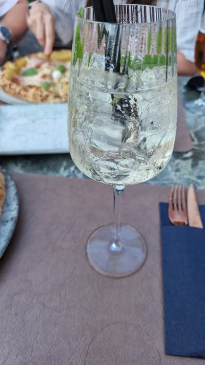 A close-up, sideways shot shows a chilled glass of white wine filled with ice and what appears to be mint leaves. The glass is condensation-covered, indicating a cool drink served in a warm environment. In the background, a pizza with toppings of ham, cheese, and fresh mint can be seen. A dark blue napkin and a rustic wooden fork are also visible on the table. The scene suggests a casual dining experience, possibly outdoors, given the lighting and the presence of greenery in the background. The image does not contain any visible text. The location context is Zadar, Croatia, which evokes a sense of a pleasant European vacation or meal.
