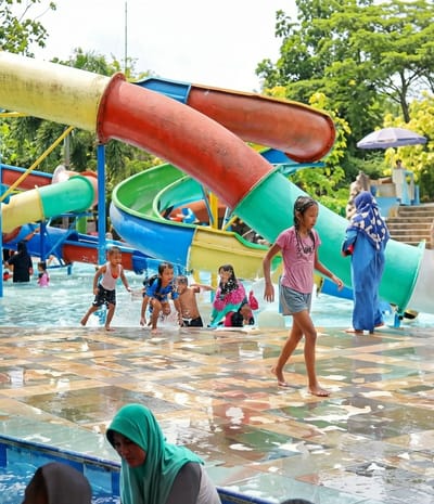 The image depicts a vibrant water park scene, likely located in Durin Simbelang, Indonesia, featuring multiple colorful water slides. A group of children are actively playing in the water, running and splashing near the slides. A young girl in a pink t-shirt and gray shorts walks barefoot on the tiled deck near the water's edge. Several women, some wearing hijabs, are present, suggesting a family-oriented setting. In the background, an adult in full Islamic attire stands near a set of stairs, further indicating a public recreational area in Indonesia. The environment is characterized by wet, tiled surfaces reflecting the water, and lush greenery in the background, possibly indicating an outdoor recreational facility.