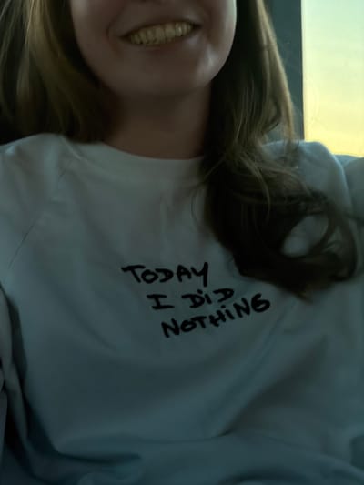 A young woman with long brown hair smiles, displaying braces on her teeth. She wears a white hooded sweatshirt with the text "Today I Did Nothing" printed on it in black letters. The background appears to be a window with a yellow-tinted light coming through it. There is no specific information or indication of the location being in the city of Loznica, Serbia.