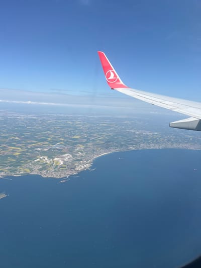 The image shows a view from an airplane window, with a portion of the wing visible in the foreground. The wing is white with a red-painted tip featuring a white emblem. Below the wing, a landscape transitions from agricultural fields to a coastline bordering a body of water. The fields appear to be a mix of green and yellow hues, suggesting different crops. Along the coastline, there is a city or town with a harbor visible, and several ships in the water. The sky is clear blue, suggesting a sunny day. The city is near the Süleymanpaşa district in Tekirdağ, Turkey.