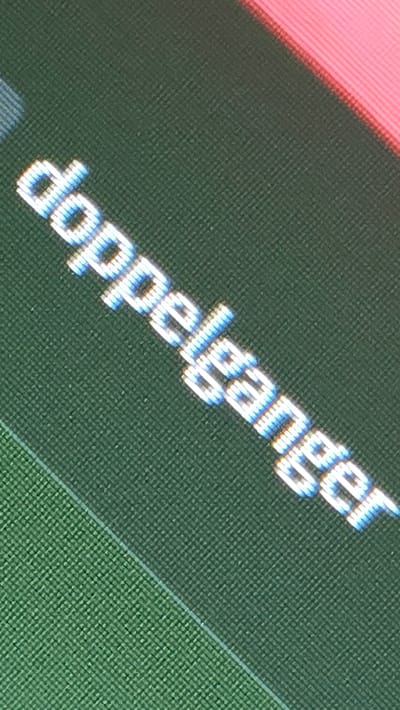 The image displays the word "doppelganger" written in a digital, pixelated font. The text is oriented diagonally from the bottom left to the top right. The letters are white with a slight blue outline and appear to be glowing. The background is a textured, dark green surface. In the upper right corner, a strip of bright pink material is visible. The image is a close-up shot, and there are no discernible geographical markers that would indicate its location in Lompoc, United States of America.