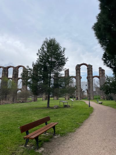 The image, oriented as a landscape, captures a serene park scene featuring ancient Roman aqueduct ruins. The impressive aqueduct, characteristic of the Acueducto de los Milagros in Mérida, Spain, stretches horizontally across the midground and background, its arches made of brick and stone standing against a cloudy sky. Birds are perched on some of the upper sections of the arches.
In the foreground, a well-maintained dirt or gravel path curves from the upper right toward the center-left. Alongside the path, a neatly mowed grassy area dominates the lower half of the frame. A dark brown wooden park bench with black metal supports is positioned in the lower right foreground, facing towards the aqueduct. Several trees are scattered throughout the grassy expanse, with a prominent evergreen tree standing in the center-left midground. Another smaller tree or bush is visible behind the bench.
Further back, near the aqueduct and path, a small, dark-framed informational display stand is visible on the grass. Through the arches of the aqueduct, a modern, white building can be seen in the distance. A single person is faintly visible walking on the path near the aqueduct in the background.
The weather appears overcast, indicated by the uniformly grey, cloudy sky, suggesting a daytime shot without direct sunlight. The scene is calm and historical, seamlessly blending ancient architecture with a natural park environment. There is no discernible text visible in the image.