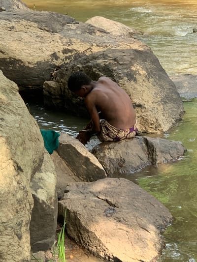 A young man is sitting on a large rock in a shallow stream. He is shirtless and wearing a pair of colorful patterned shorts. He is cupping his hands in the water, possibly washing them. He is sitting near the bank of the stream, surrounded by large rocks. The stream is flowing gently, and the water is a light green color. There is a green cloth lying on the ground nearby. The location is a small stream in the Egbeda area of Lagos, Nigeria, specifically at the coordinates 7.119° N, 5.362° E.