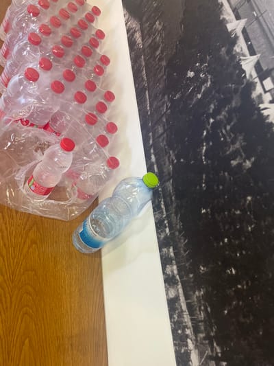 The scene is located within the Brener Regional Council, Israel. The image depicts a close-up, high-angle view of several objects arranged on a multi-textured surface. In the upper left and central portions, a large multi-pack of approximately 36 clear plastic water bottles with red screw caps is visible. Most of these bottles appear to be empty or nearly empty, still enclosed within their transparent plastic shrink wrap packaging. One bottle from this pack, partially filled with liquid, is seen detached from the main bundle, resting below it. To the right of the packaged bottles, a single, clear plastic water bottle with a neon green cap lies on its side, partially filled with liquid, featuring a blue and white label.
The environment consists of a light brown wooden floor with visible grain occupying the lower left quadrant of the image. A distinct white, flat surface acts as a linear divider, separating the wooden floor from a large, vertical black and white photographic print or poster on the right side. This print displays a detailed, textured image, possibly of trees or dense foliage. No individuals are present, and no active interactions are depicted.