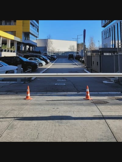 In Bielawska Wieś, Poland, a modern parking area is depicted under a clear blue sky. Two orange traffic cones are positioned on a grey paved surface, one to the left and one to the right, casting distinct shadows. A white horizontal barrier stretches across the mid-ground. Beyond the barrier, a row of parked cars is visible, including black SUVs and a silver hatchback. In the background, contemporary buildings with yellow and blue facades stand, along with a white rectangular structure and trees. A black Jeep is also present in the parking area. The scene appears to be a secure, organized, and well-maintained urban space.