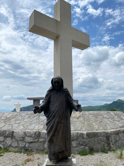 The image depicts an outdoor religious site in Benkalj, Montenegro, under a partially cloudy sky. In the immediate foreground, a large, dark-colored statue of a hooded figure, likely a saint or monastic, stands with arms slightly outstretched. One hand clutches a rosary. The statue is made of a dark material, possibly bronze or stone, showing texture and drapery in its robe.
Behind and to the left of this statue, a monumental, light-colored concrete cross rises against the sky, its horizontal arm also visible, dominating a significant portion of the scene. Further in the background, a smaller, white statue resembling Christ the Redeemer, with outstretched arms, is positioned on a distant elevation.
The ground around the statues consists of gravel and sparse green vegetation. A sturdy stone wall, constructed from irregular grey and brown stones, runs across the midground. Beyond these structures, a range of mountains can be seen, indicating a high-altitude or scenic location.
The time of day appears to be daytime, possibly late morning or early afternoon, given the natural lighting. The weather is fair, with patches of blue sky visible between fluffy white clouds. No specific activity or event is taking place, as the image captures static religious monuments. There is no visible text in the image. The scene conveys a sense of solemnity and peace.