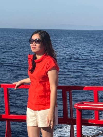 An East Asian woman stands on a boat, leaning on a red railing. She is wearing a red short-sleeved top with a ruffled collar and off-white shorts. She has on large, dark sunglasses and her long, dark hair is down. The background is a vast expanse of blue water under a clear sky, with distant land visible on the horizon. The scene suggests a travel or leisure activity on the water, possibly a ferry or cruise. The specific location within Dong Son, Vietnam, cannot be determined from the visual information present in the image.